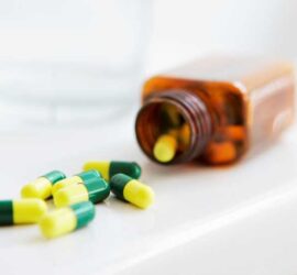 Green and yellow capsules spilling from a brown pill bottle on a white surface.