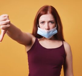 Woman with mask down giving a thumbs down against orange background.
