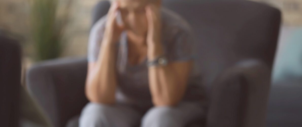 An elderly woman sitting on a couch looking distressed with a phone on a table.
