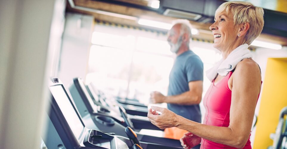 Older adults exercising on treadmills, smiling brightly.