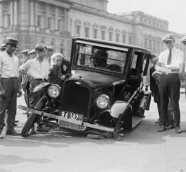 A vintage car stuck in a large pothole on a city street with onlookers gathered.