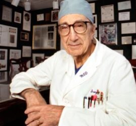 Doctor in office with wall of certificates.