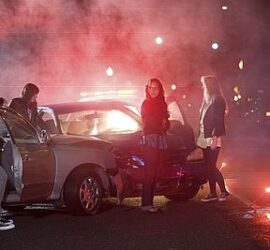 Three people posing near a vintage car with dramatic red lighting and fog.
