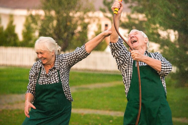 Elderly couple joyfully playing with garden hose.